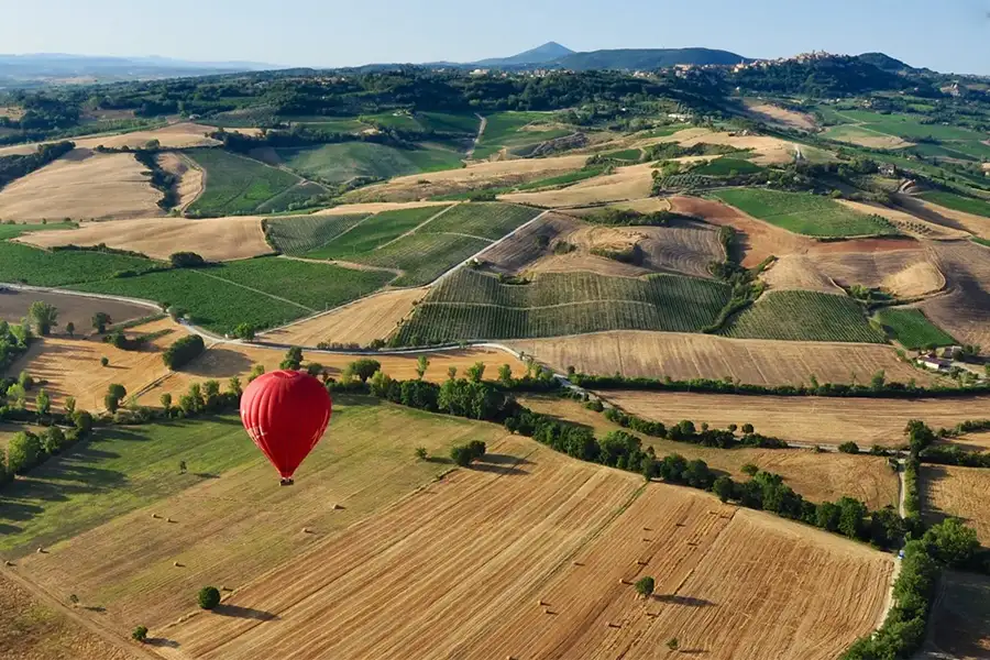 Red hot air balloon flying over the hills and cultivated fields of Tuscany, with a panoramic view of the rural landscape