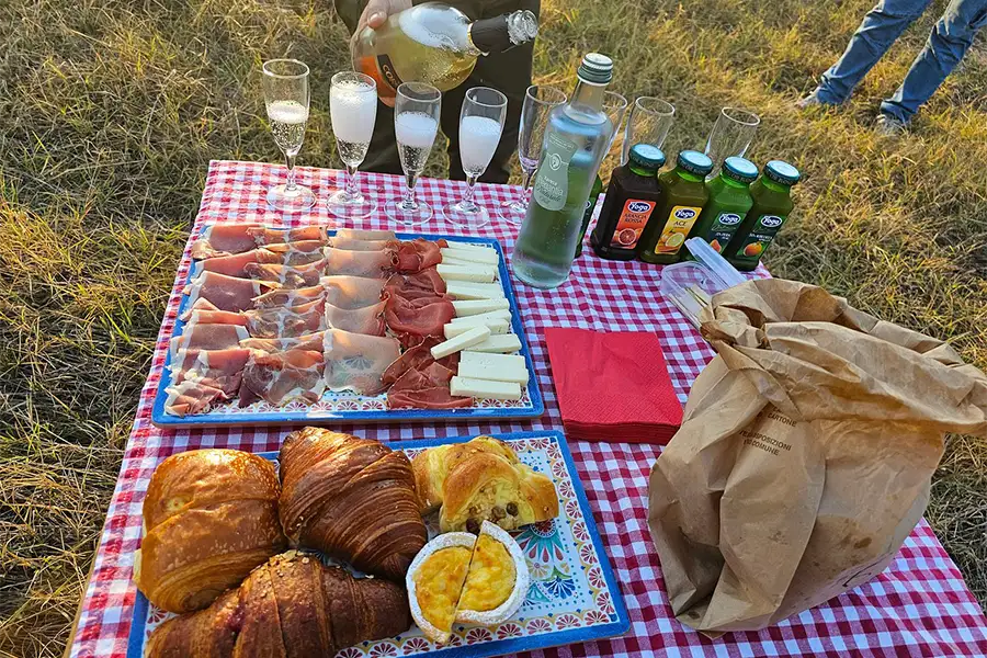 Café da manhã ao ar livre com frios, queijos, croissants, sucos e espumante sobre toalha xadrez vermelha, após passeio de balão na Toscana