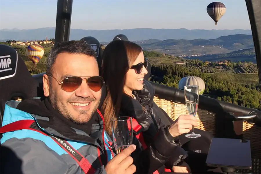 Smiling couple enjoying a hot air balloon ride over the countryside with glasses of champagne, surrounded by scenic hills and other balloons in the background.